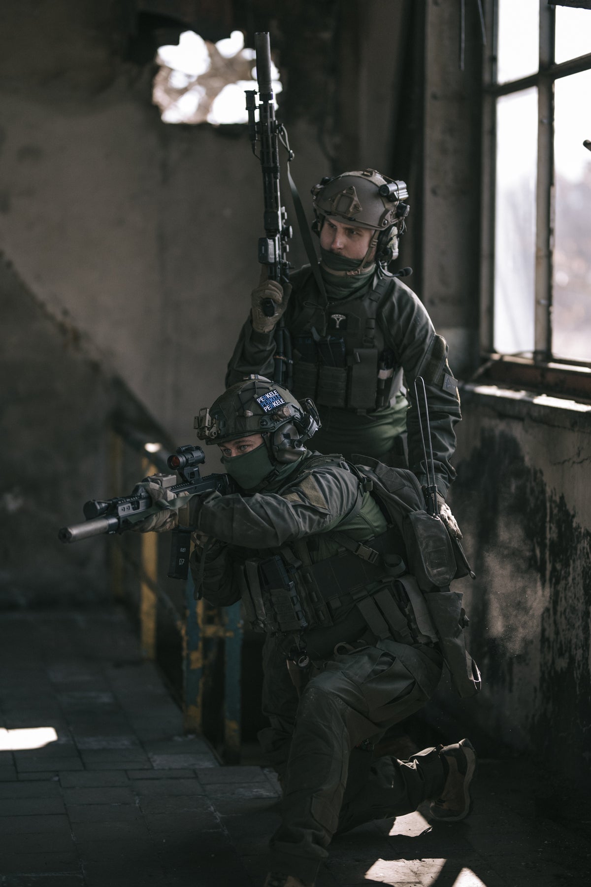 Two soldiers in tactical gear inside a dimly lit building.