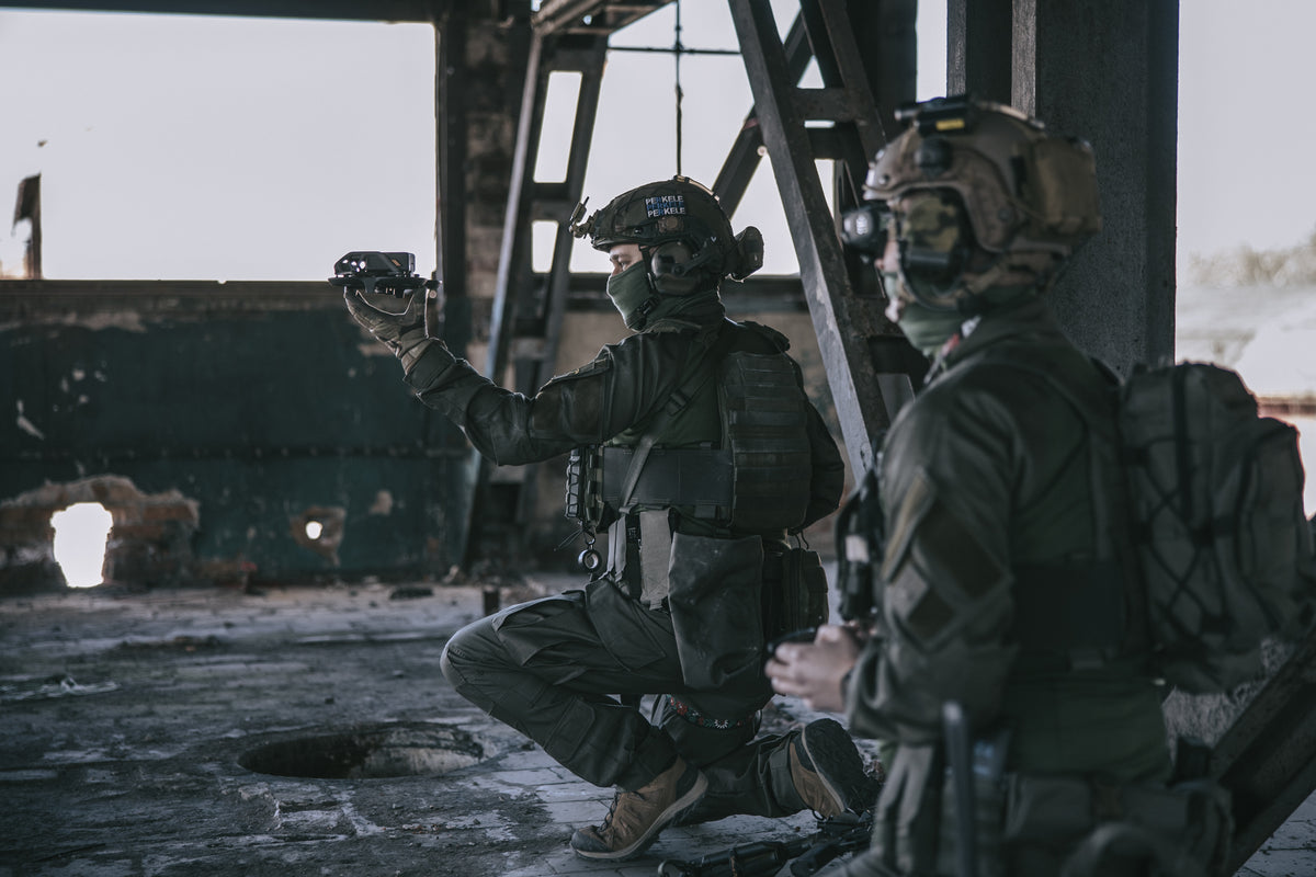 Two soldiers in tactical gear inside a dilapidated building, one of whom is holding a drone in his hands, ready to launch it.