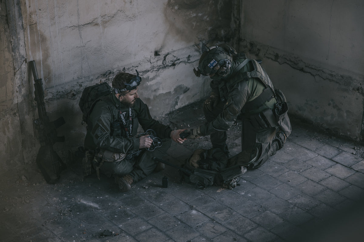 Zwei Soldaten in taktischer Ausrüstung sitzen auf einem Betonboden in einem schwach beleuchteten Raum.