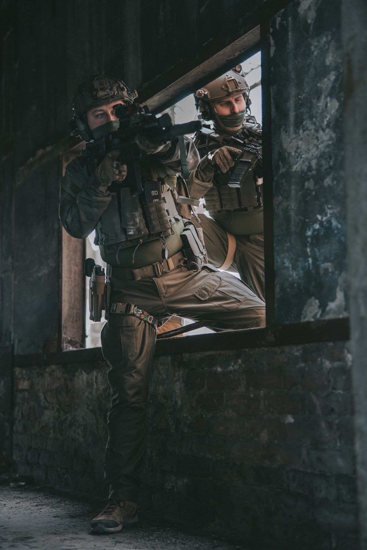 Two soldiers in tactical gear climbing through a window in an industrial area