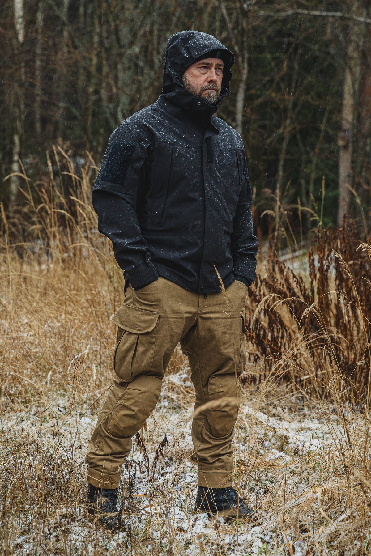 Man wearing a black jacket and tan pants standing in a forest with snow on the ground.