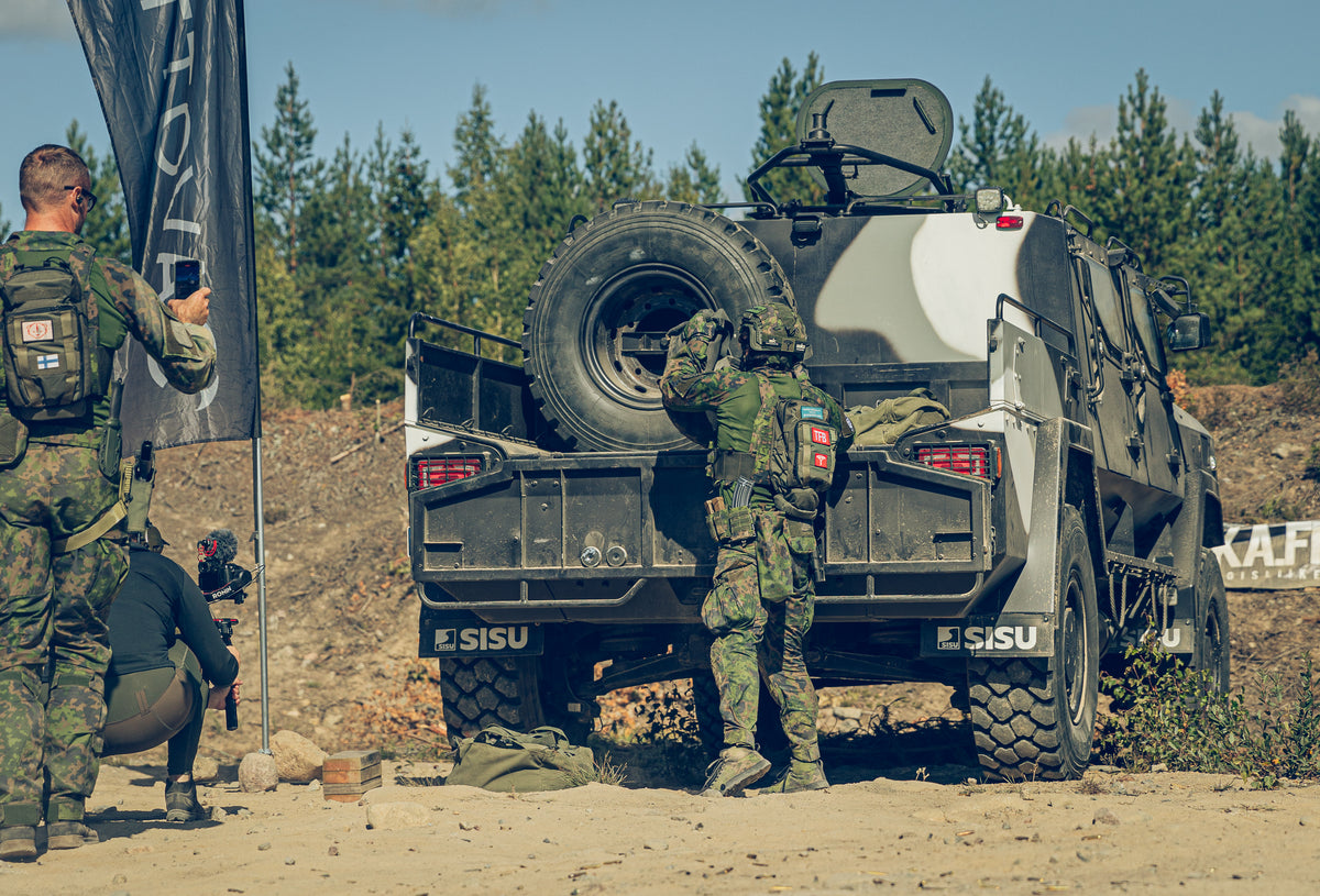 Two soldiers in camouflage next to a military vehicle with 'SISU' branding, outdoors.