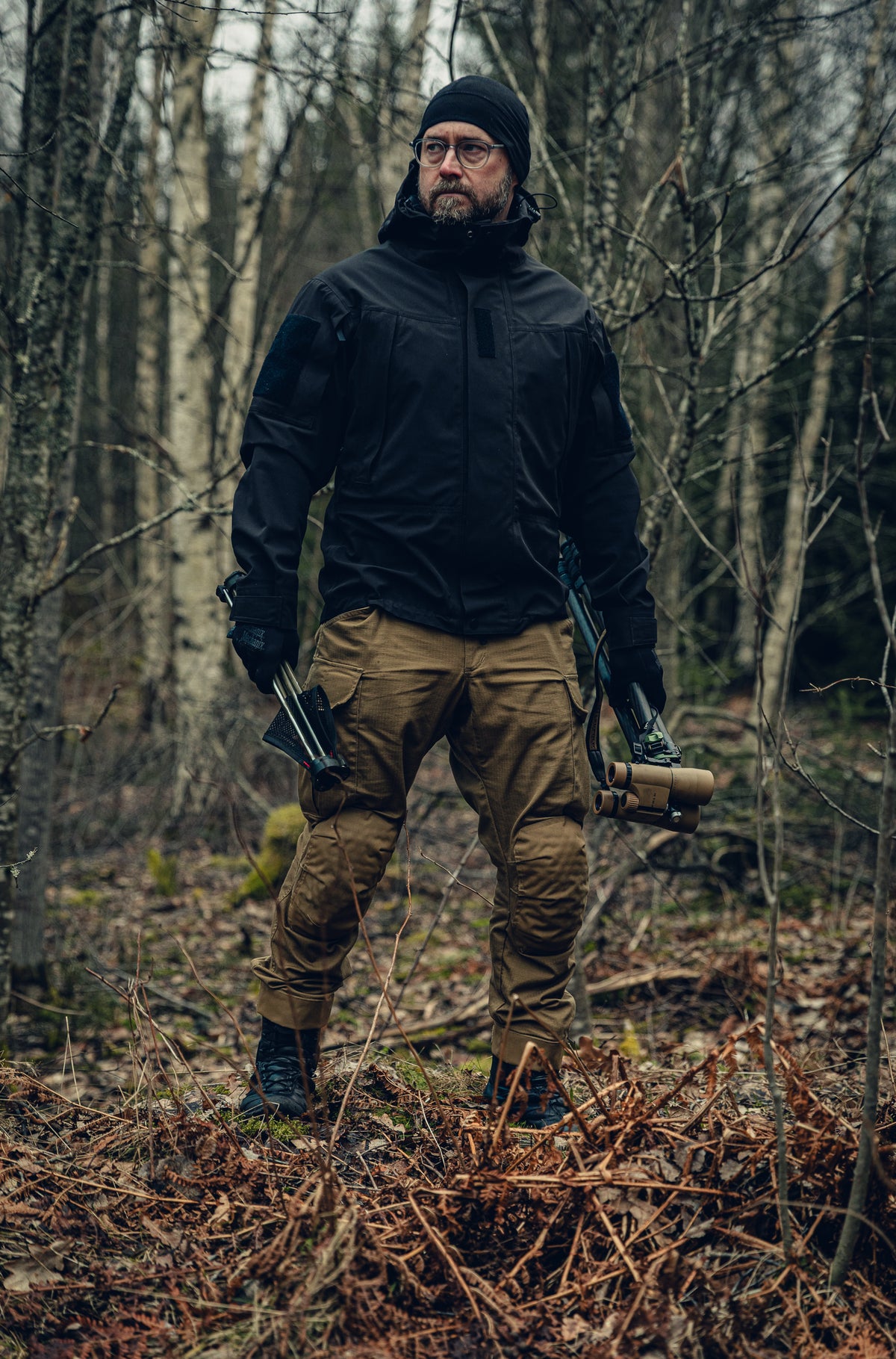 A man standing in a birch wood forest looking to the left. He is wearing a black jacket and beanie and light brown cargo pants and holding binoculars in one hand and a small foldable stool in the other hand.