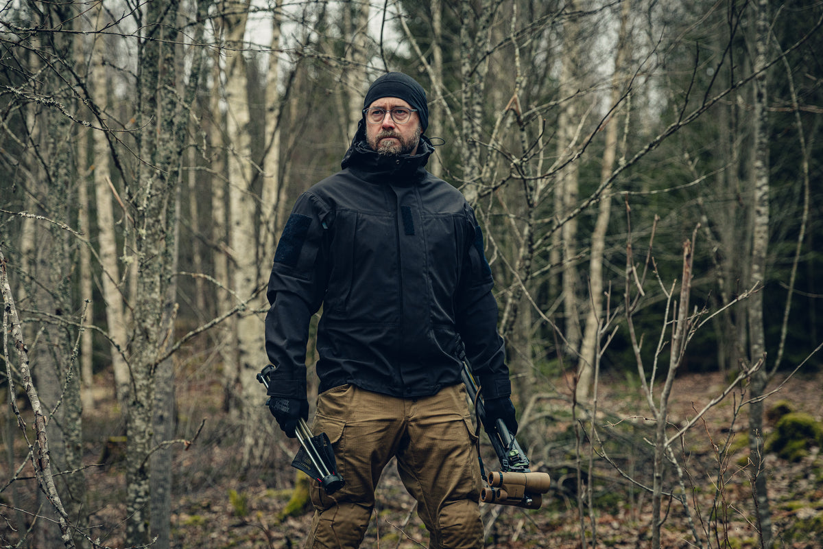 Ein Mann steht in einem Birkenwald und schaut nach links. Er trägt eine schwarze Jacke und Mütze sowie hellbraune Cargohosen und hält in einer Hand ein Fernglas und in der anderen einen kleinen klappbaren Hocker.