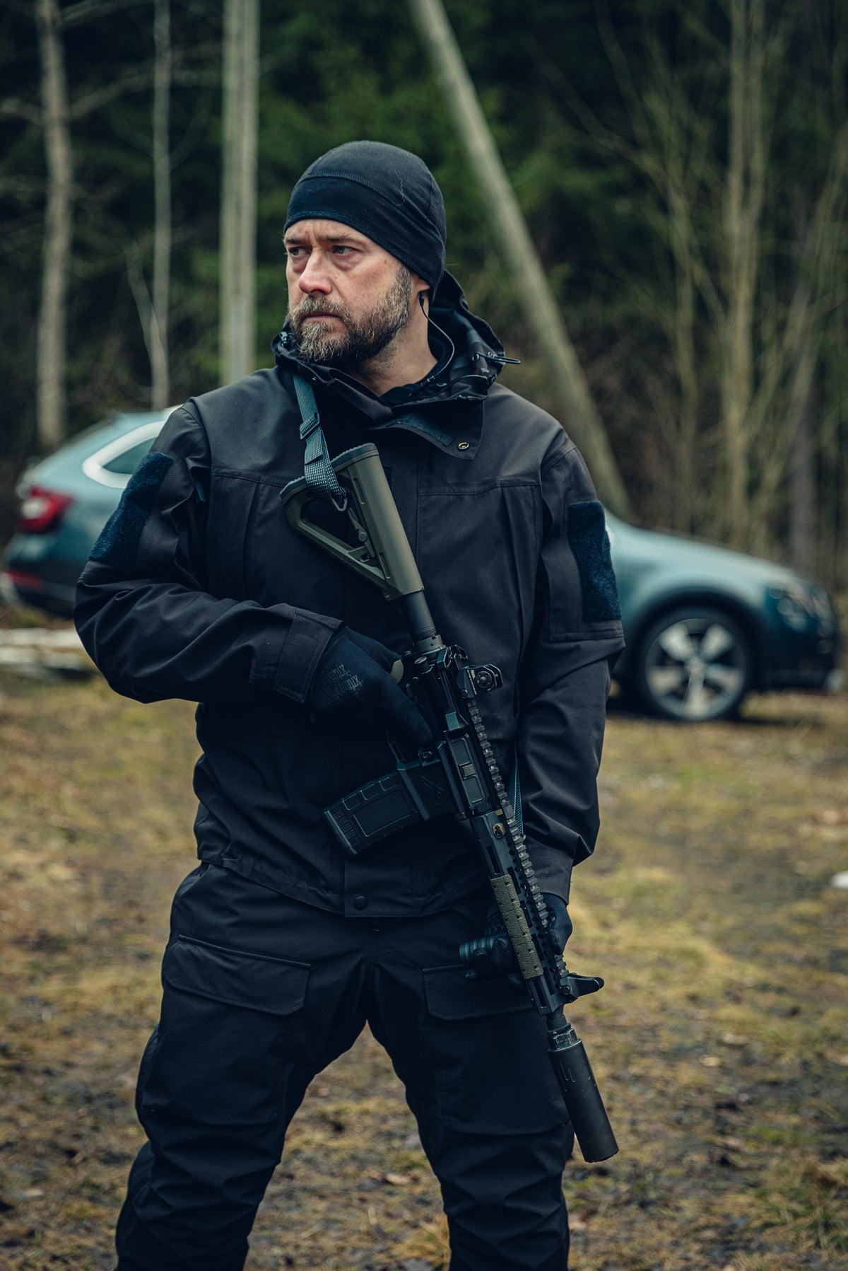 Ein Mann steht vor einem graublauen Auto, trägt ein Sturmgewehr und schaut nach links. Er trägt eine schwarze Hose, eine schwarze Jacke und eine schwarze Mütze, und aus seinem Ohr kommt ein Kabel.