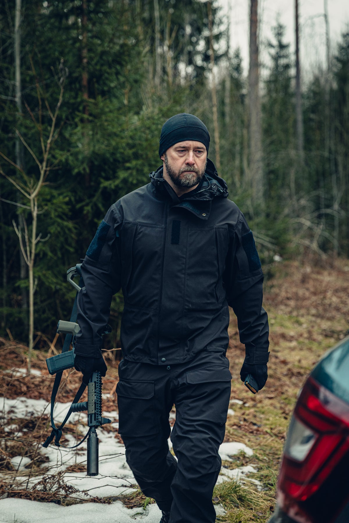 Ein Mann hält ein Gewehr in der rechten Hand und geht auf den Kofferraum eines Autos zu. Er trägt eine schwarze Jacke, eine schwarze Mütze und eine schwarze Hose.