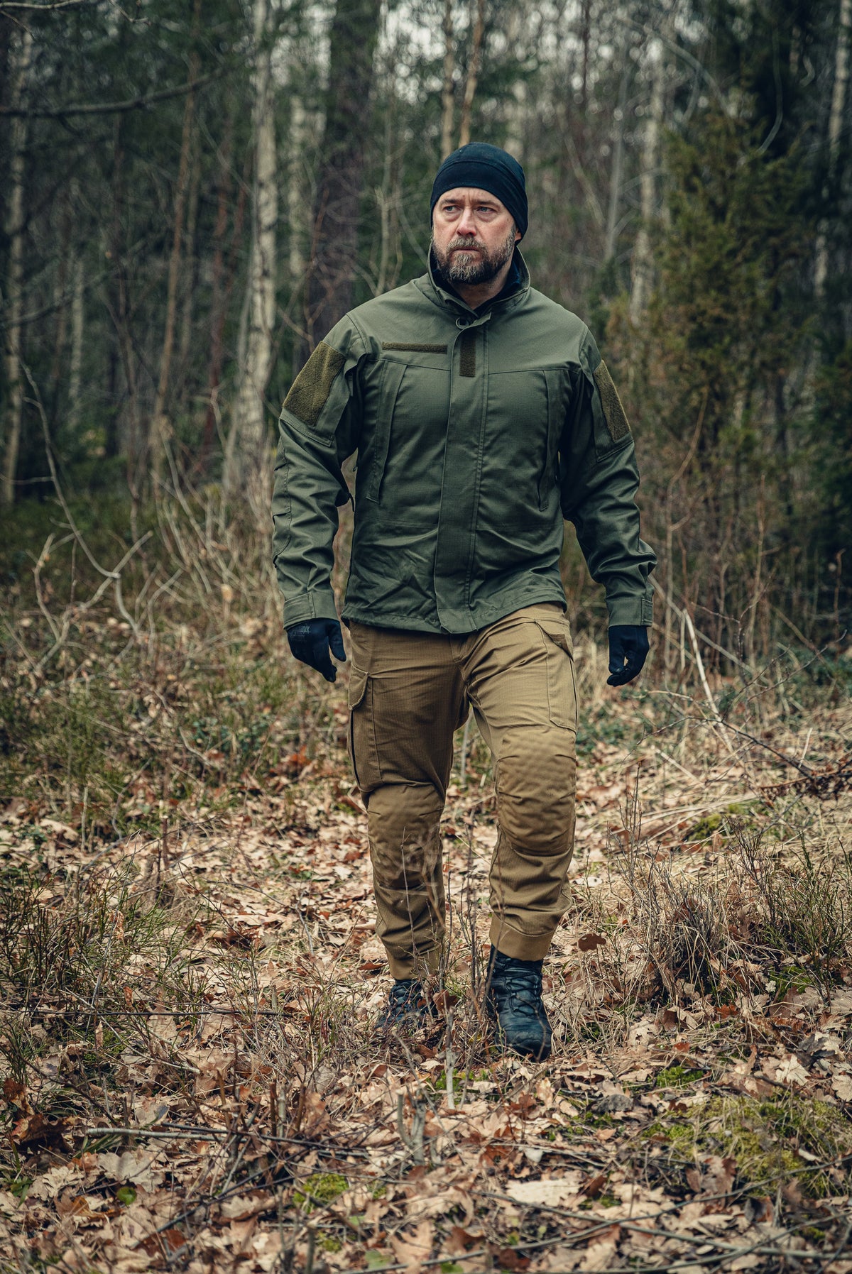 A bearded man walking in a forest looking slightly to the left. He is wearing a green jacket with a snug fit, a black beanie and gloves and light brown cargo pants.