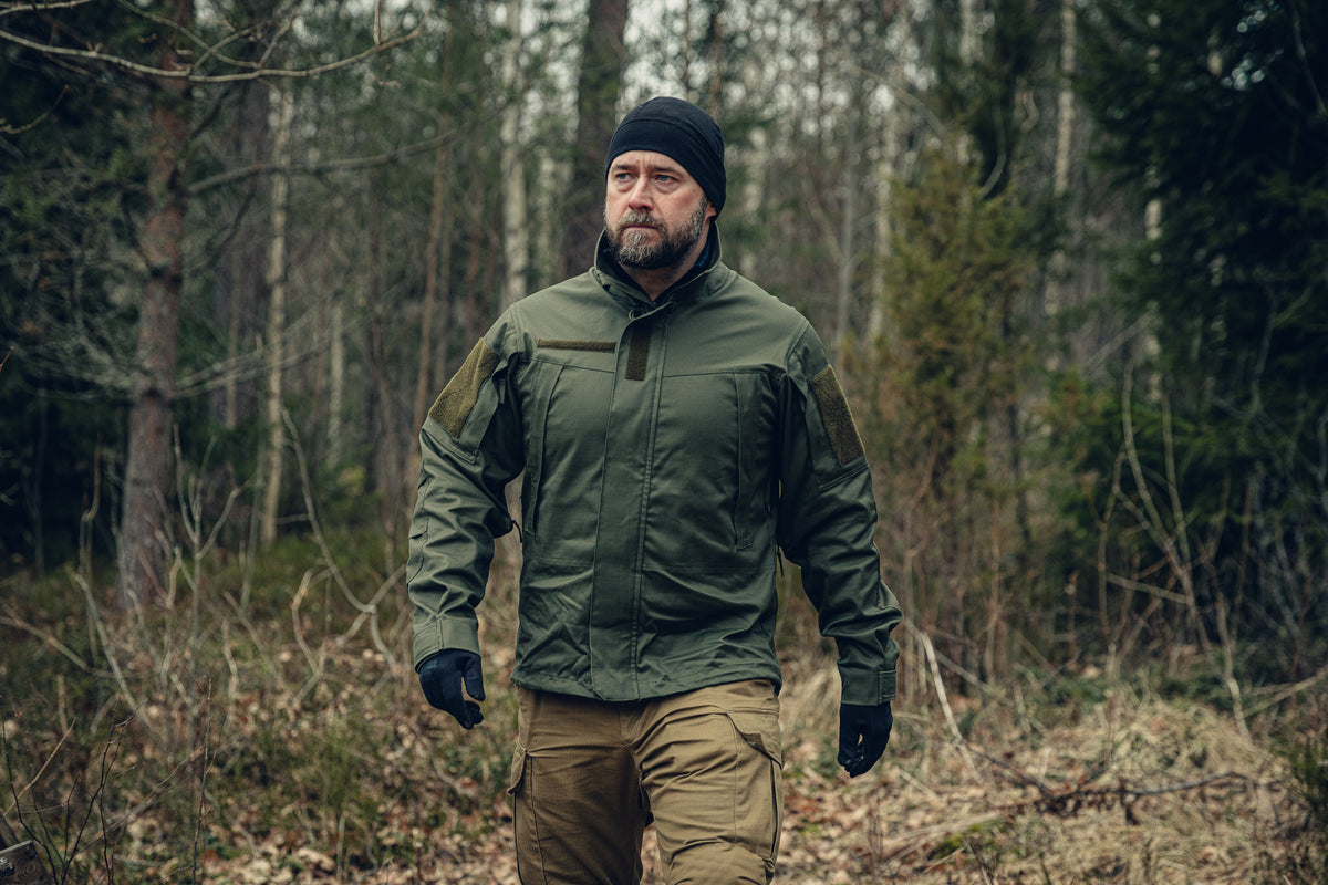 Ein bärtiger Mann geht durch einen Wald und blickt leicht nach links. Er trägt eine grüne, eng anliegende Jacke, eine schwarze Mütze und Handschuhe sowie hellbraune Hosen.
