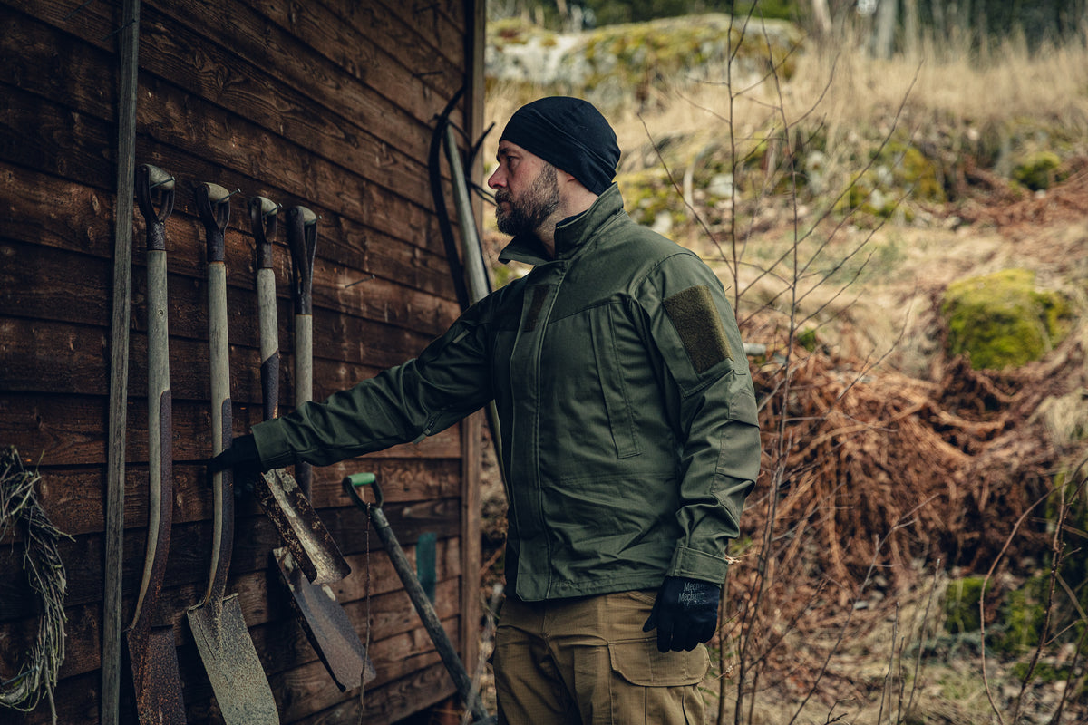 A bearded man puttin a shovel to a wooden wall with other shovels hanging off of it. He is wearing a green jacket with a snug fit, a black beanie and gloves and light brown pants.