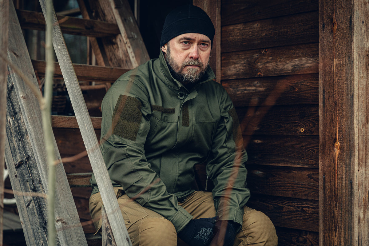 Ein düster wirkender Mann sitzt auf einer Holztreppe vor einer alten Holzhütte. Er trägt eine grüne, eng anliegende Jacke, eine schwarze Mütze und Handschuhe sowie hellbraune Cargohosen.