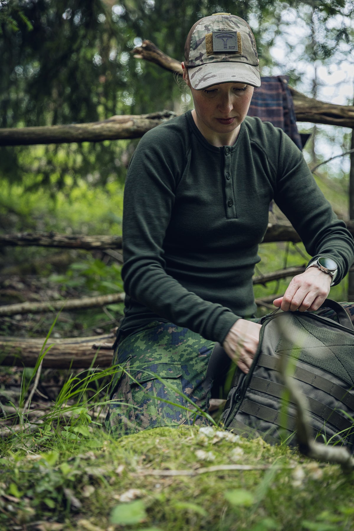 A woman kneeling outdoors in a wooded setting, wearing a dark olive long-sleeve henley shirt, camouflage pants, and a camouflage cap while packing or organizing a dark tactical-style bag.