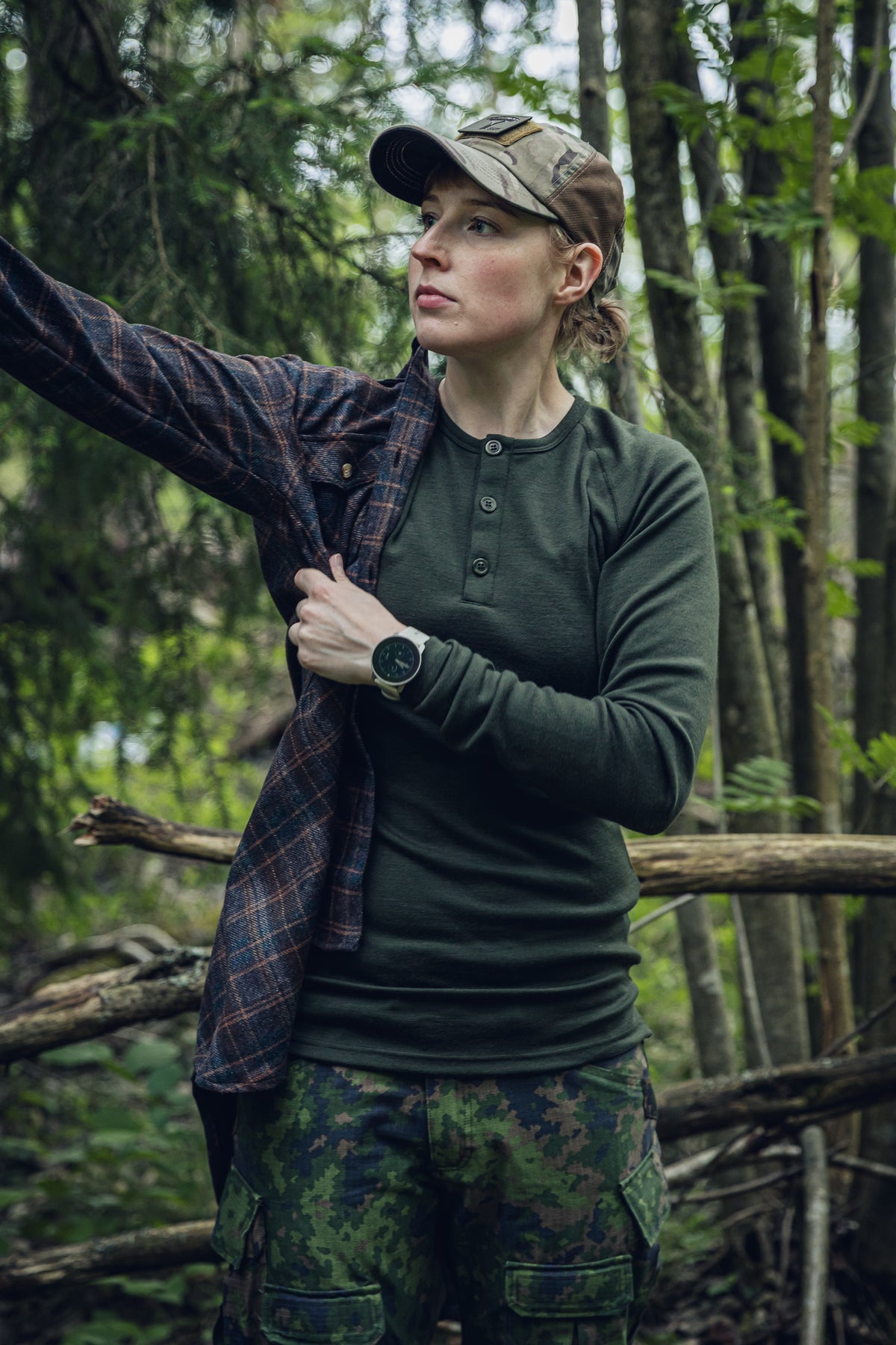 A woman in a forest wearing a dark olive green long-sleeve henley shirt, camouflage pants, a camouflage cap, and a dark plaid overshirt.