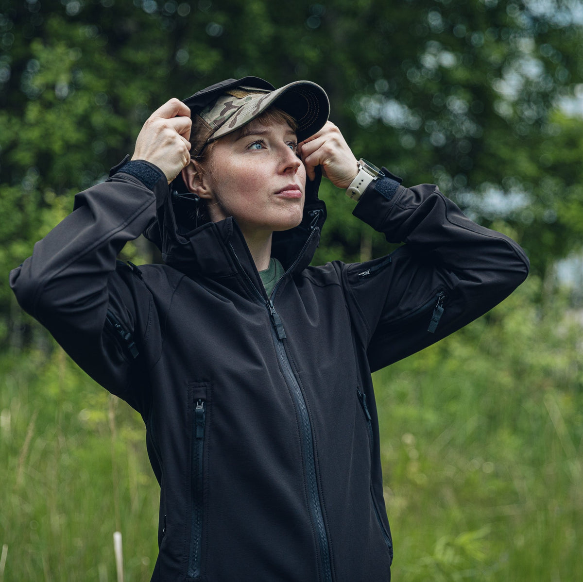 A woman wearing a black outdoor  jacket and a camouflage cap stands in a grassy field with trees in the background, adjusting her hood.