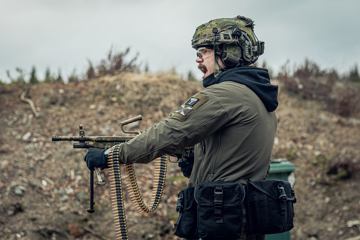 A person in tactical gear, including a camouflage helmet, olive drab jacket, and black utility pouches, holding a submachine gun with a mounted belt loader in an outdoor, rugged terrain setting.