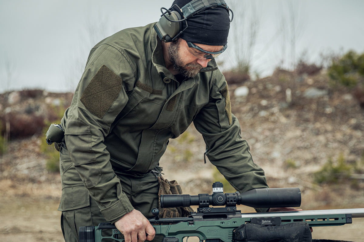 Man in tactical gear adjusting a rifle with a scope in an outdoor setting