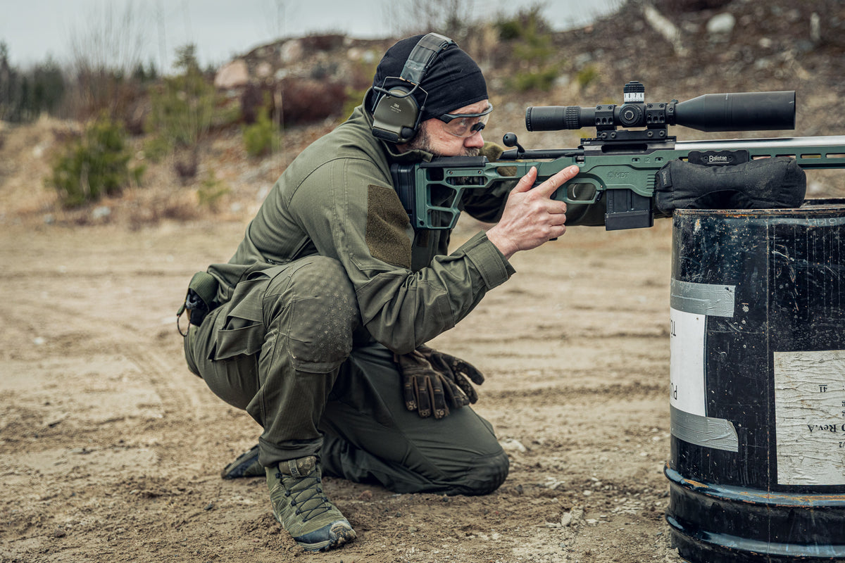 Person in military gear with a rifle, crouching behind a barrel in an outdoor setting