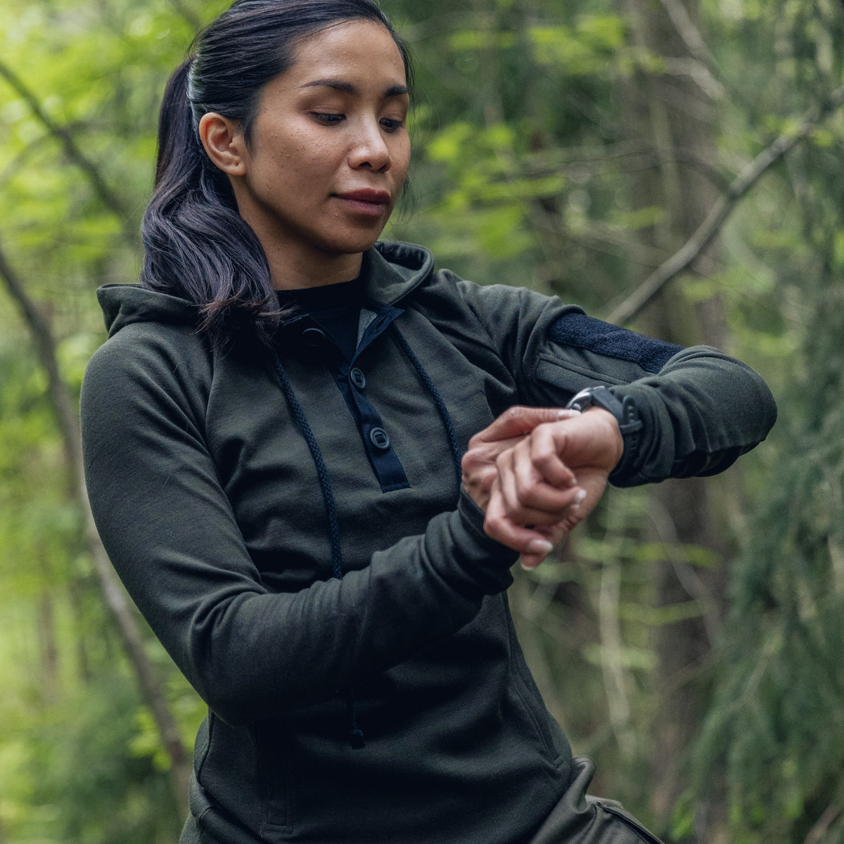 A woman in a forest setting wearing a dark olive green hooded long-sleeve shirt and looking down at her watch.