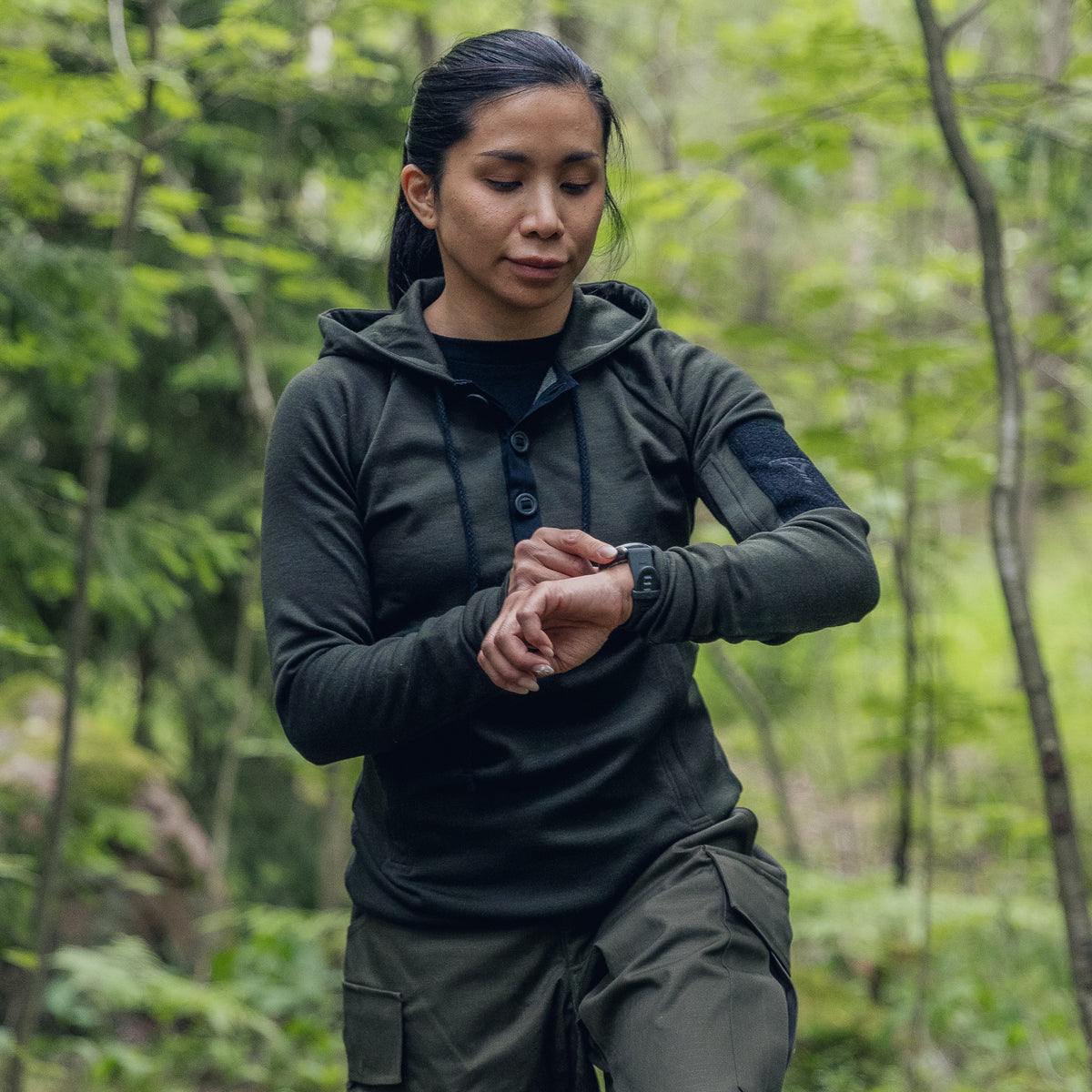 A woman wearing a dark olive green tactical hoodie and matching cargo pants stands in a forest, looking down at her wrist watch.