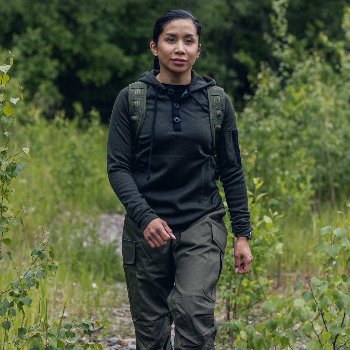 A woman wearing an olive green tactical hoodie, matching cargo pants, and a backpack walks along a path surrounded by green foliage.