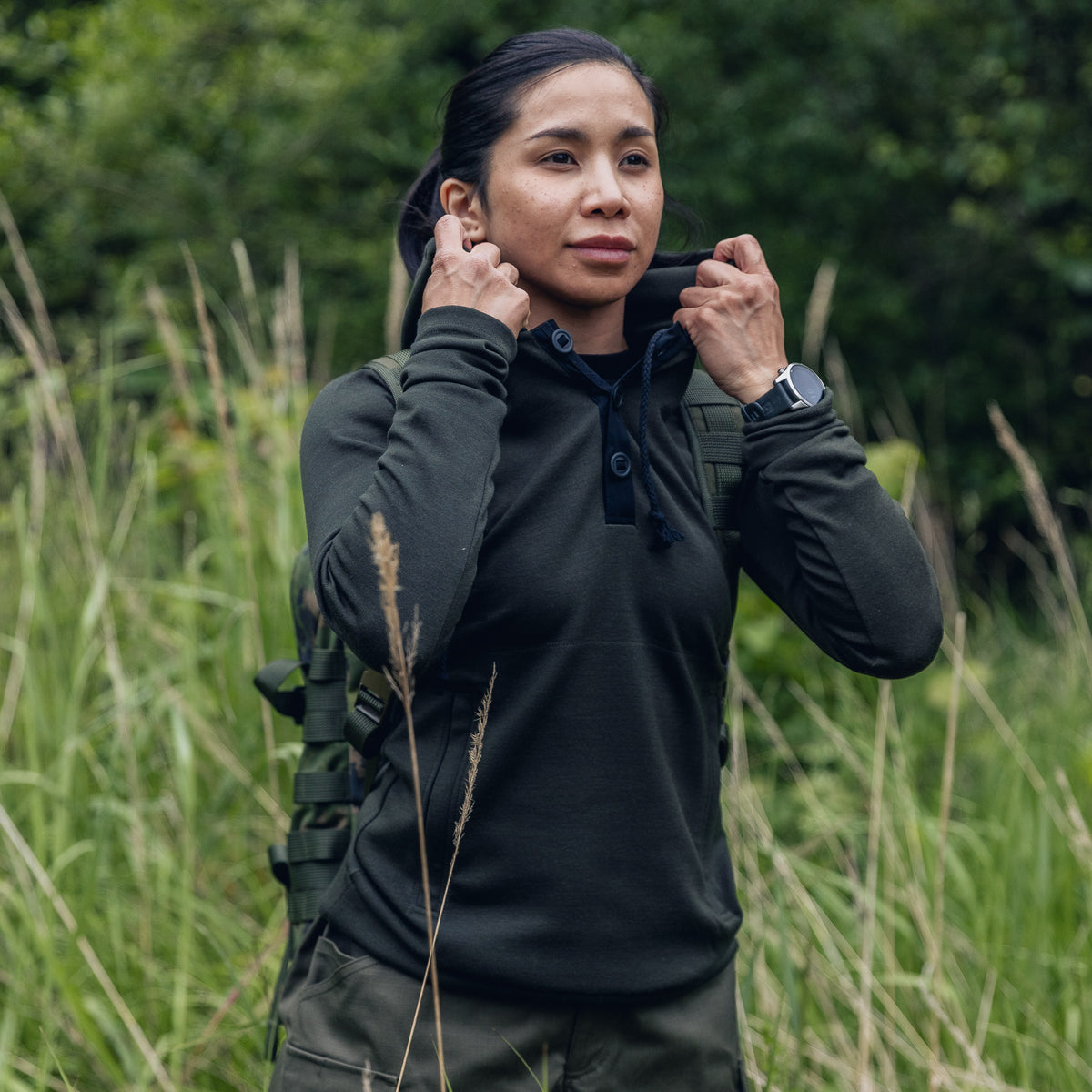 A woman outdoors in tall grass wearing a dark olive green long-sleeve pullover top and matching green outdoor gear, adjusting the collar of her shirt.