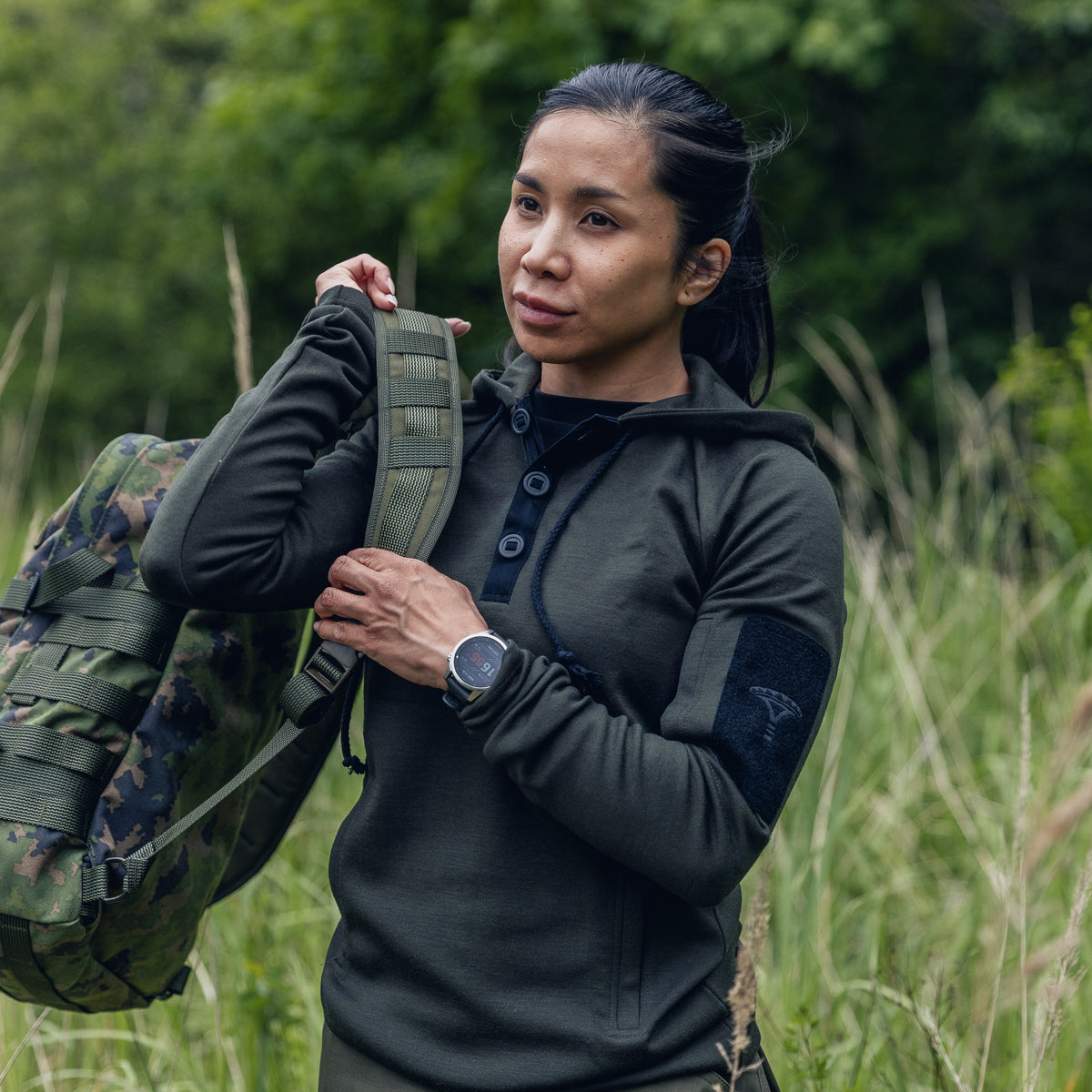 A woman outdoors in tall grass wearing a dark olive green hooded long-sleeve top and carrying a camouflage backpack.