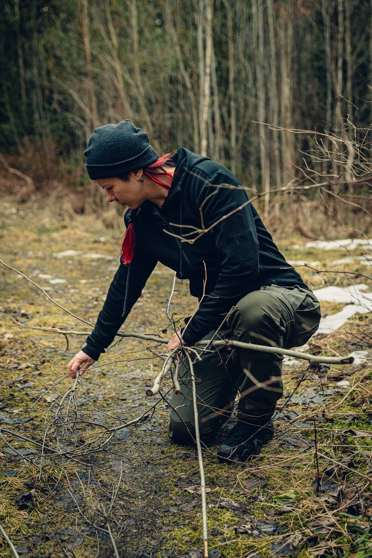 A woman kneeling on the ground gathering some sticks. She is wearing green cargo pants, a black hoodie and a grey beanie.