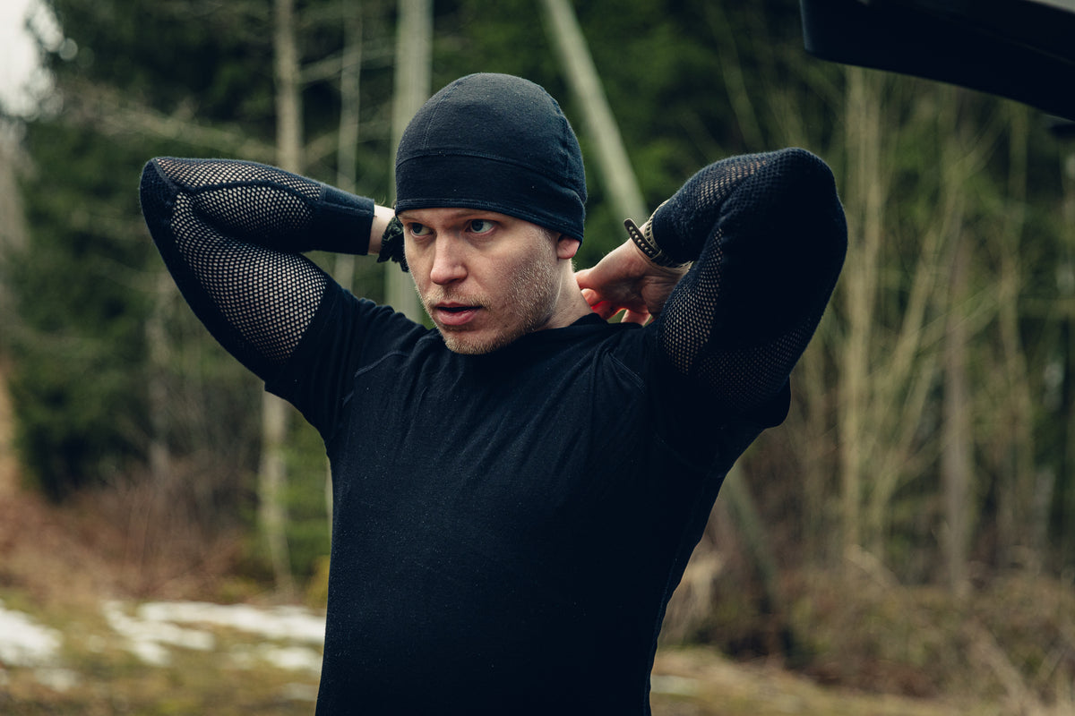 Ein junger blonder Mann trägt ein schwarzes T-Shirt, eine schwarze Mütze und ein schwarzes langärmliges Mesh-Shirt. Er scheint das Rückenteil seines Shirts zu richten. Im Hintergrund ist ein leicht verschneiter Wald zu sehen.