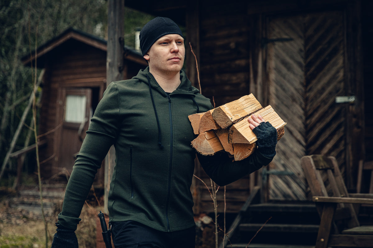 A man walking through a yard holding pieces of firewood. He is wearing a black beanie, green hoodie, fingerless gloves, and there is a knife on his belt. There are some old wooden cabins in the background.