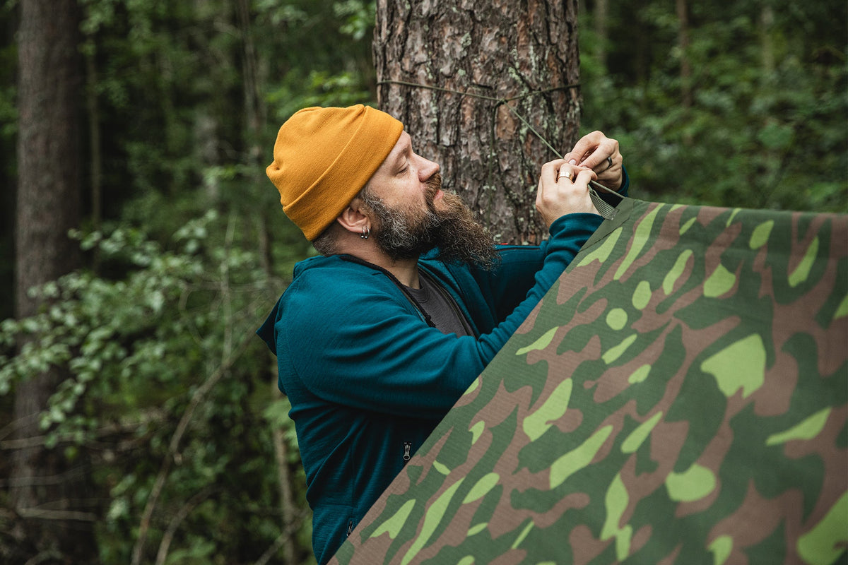 A man setting up a M91 tarp in the woods. He is wearing a yellow watch cap and teal merino wool hoodie.