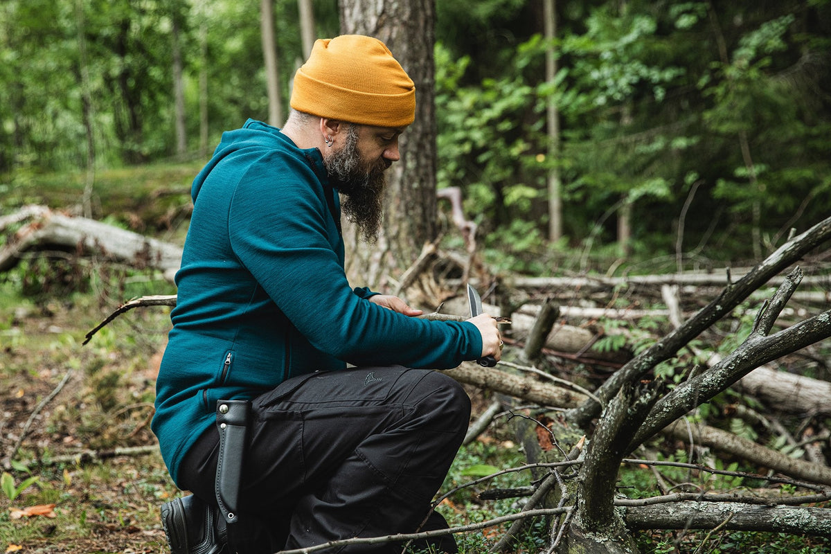 A man is crouching in the forest in a teal merino wool hoodie and yellow watch cap. He is whittling a stick.