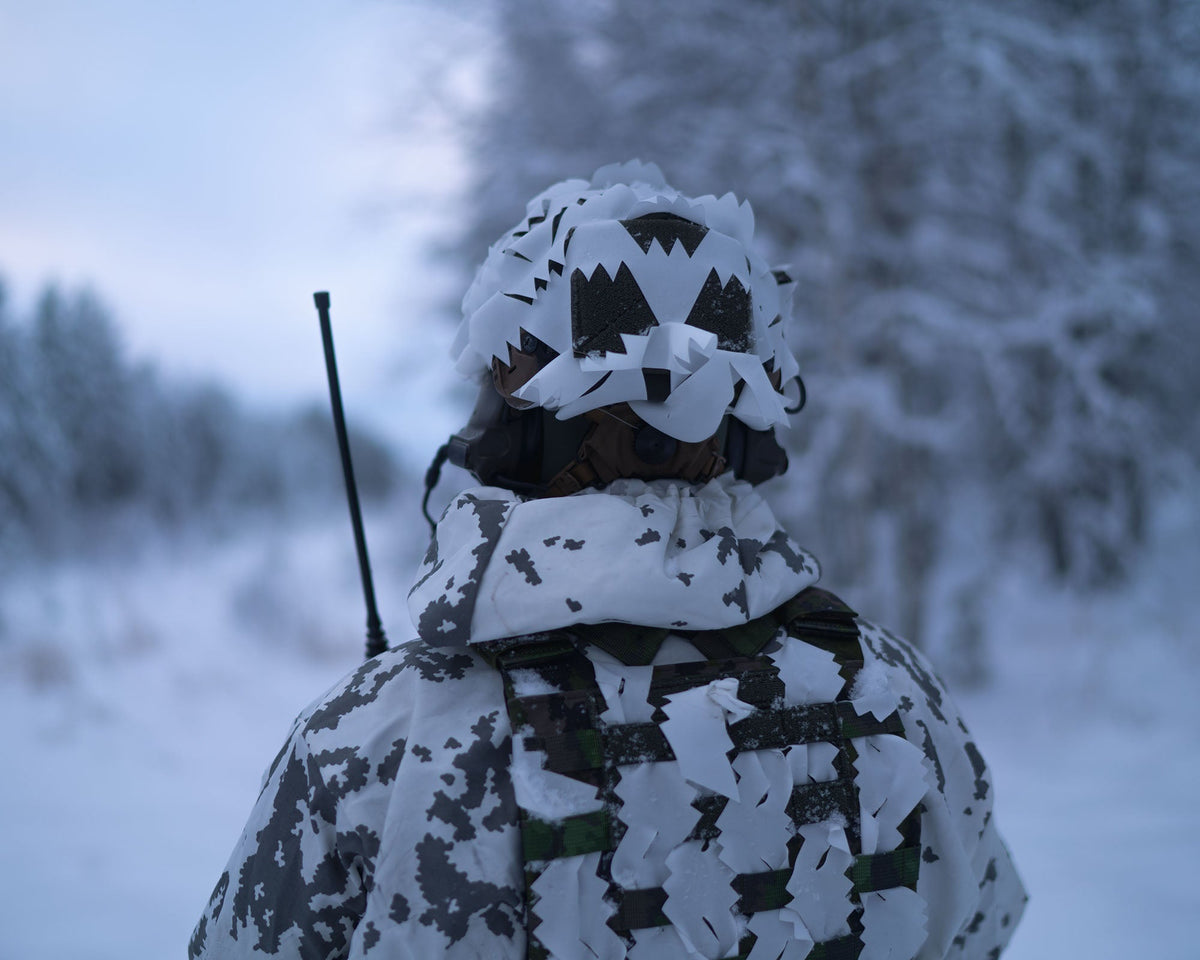 A soldier at the edge of a snowy forest wearing a helmet with white Savotta Helmet Camo Scrim.