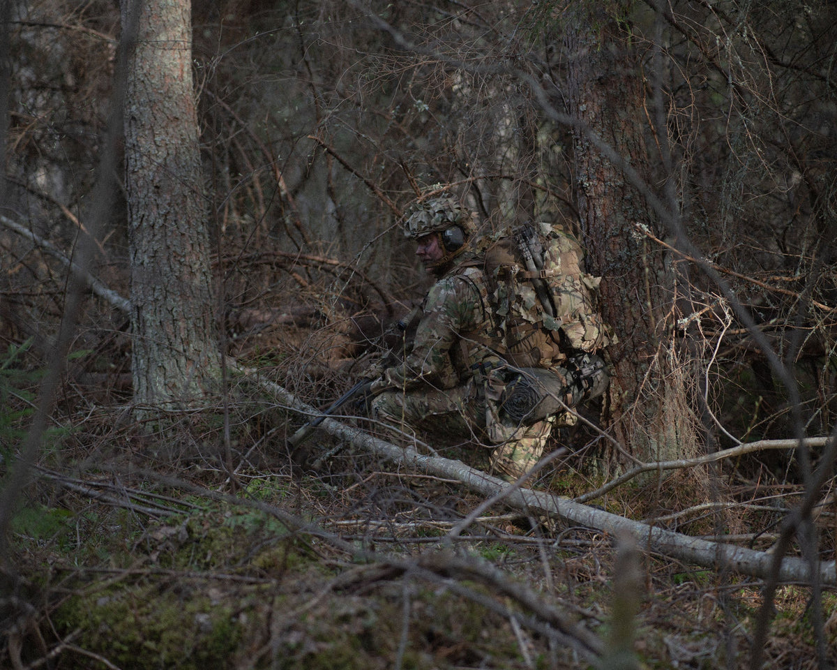 A crouched soldier in a forest wearing a helmet with MultiCam Savotta Helmet Camo Scrim.