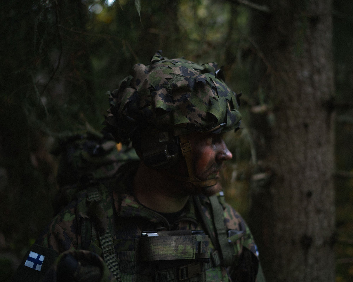 A soldier in a dark forest wearing a helmet with Finnish M05 Savotta Helmet Camo Scrim.