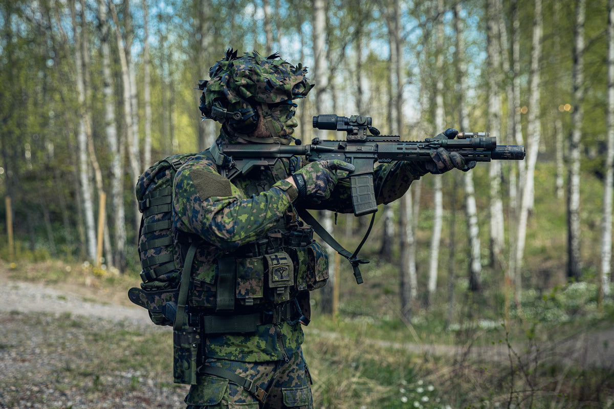 A soldier in a Finnish M05 camouflage uniform and helmet aims a black tactical rifle with a scope in a wooded outdoor setting.