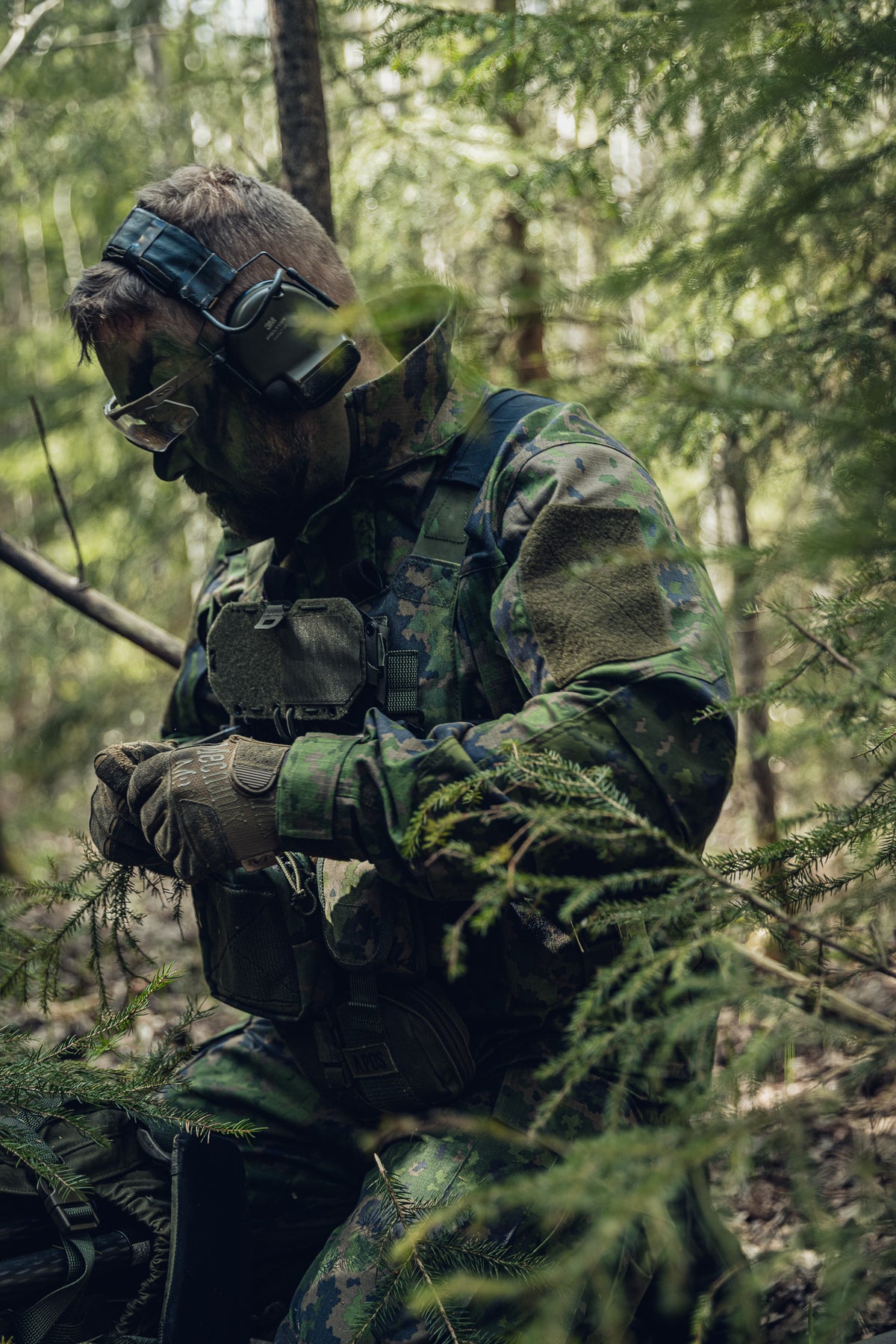 Ein Soldat in finnischer M05-Tarnung kniet auf dem Boden in einem Fichtenwald.