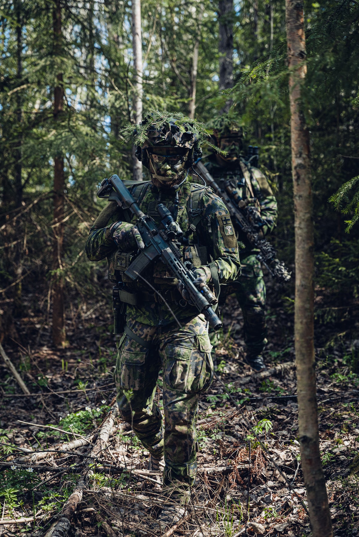 Two soldiers in full Finnish M05 camouflage military gear, including helmets, goggles, and tactical rifles, walking through a dense forest.