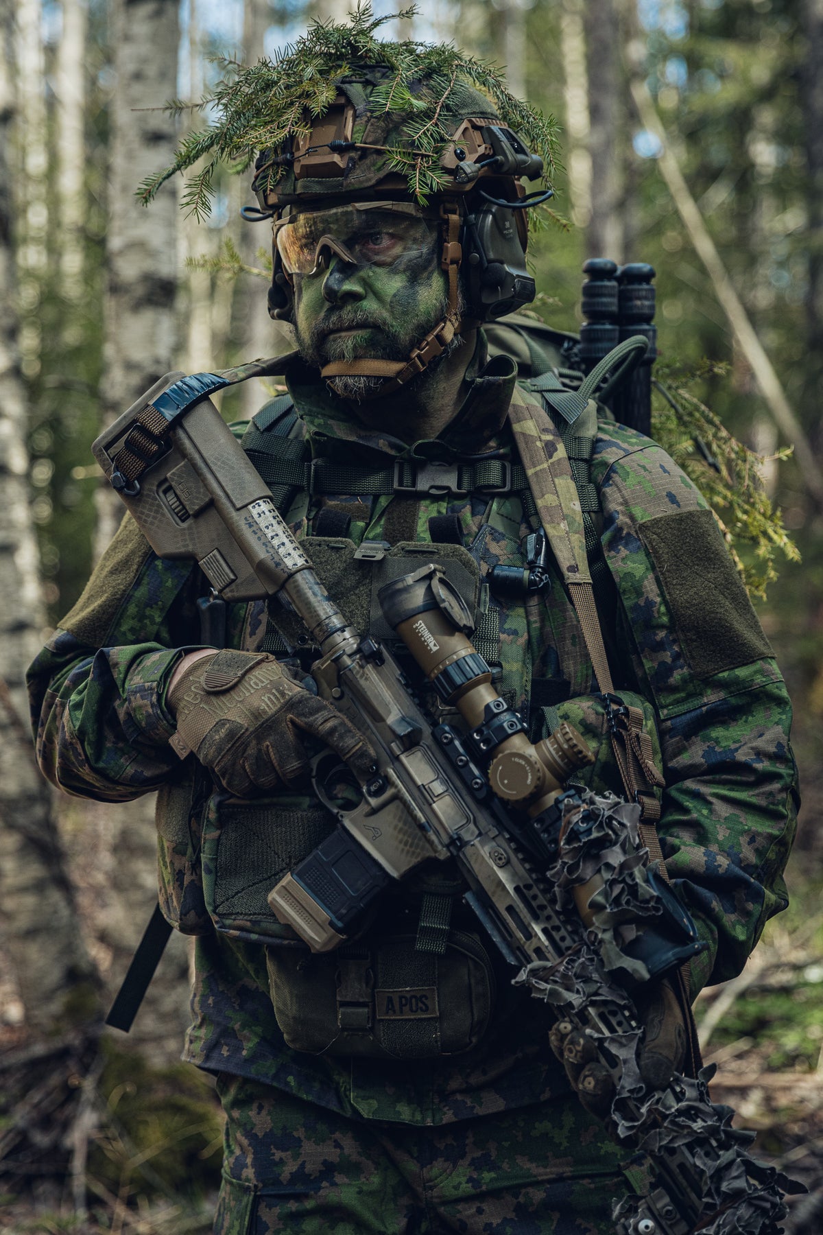 Ein Soldat in vollständiger finnischer M05-Tarn-Militär­ausrüstung, einschließlich eines mit Laub getarnten Helms, Gesichtsbemalung und eines Gewehrs mit Zielfernrohr, steht in einem Wald.