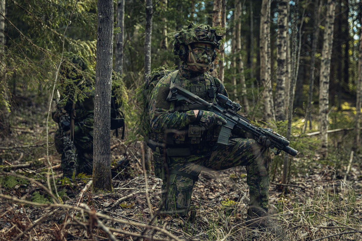 Two soldiers in full Finnish M05 camouflage military gear, crouching in a dense forest while carrying rifles.