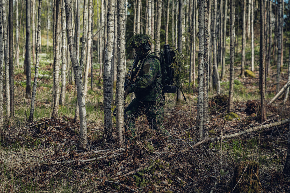 A man in Finnish M05 camo gear holding a rifle moving through a birch forest.