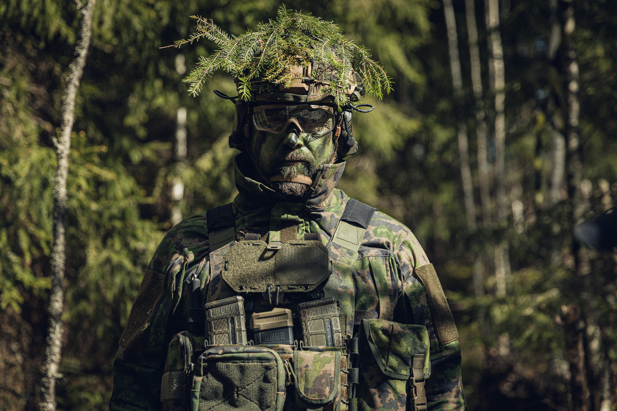A person in full military-style camouflage gear stands in a forest, wearing a helmet with leafy camouflage netting, protective goggles, and a plate carrier with pouches.