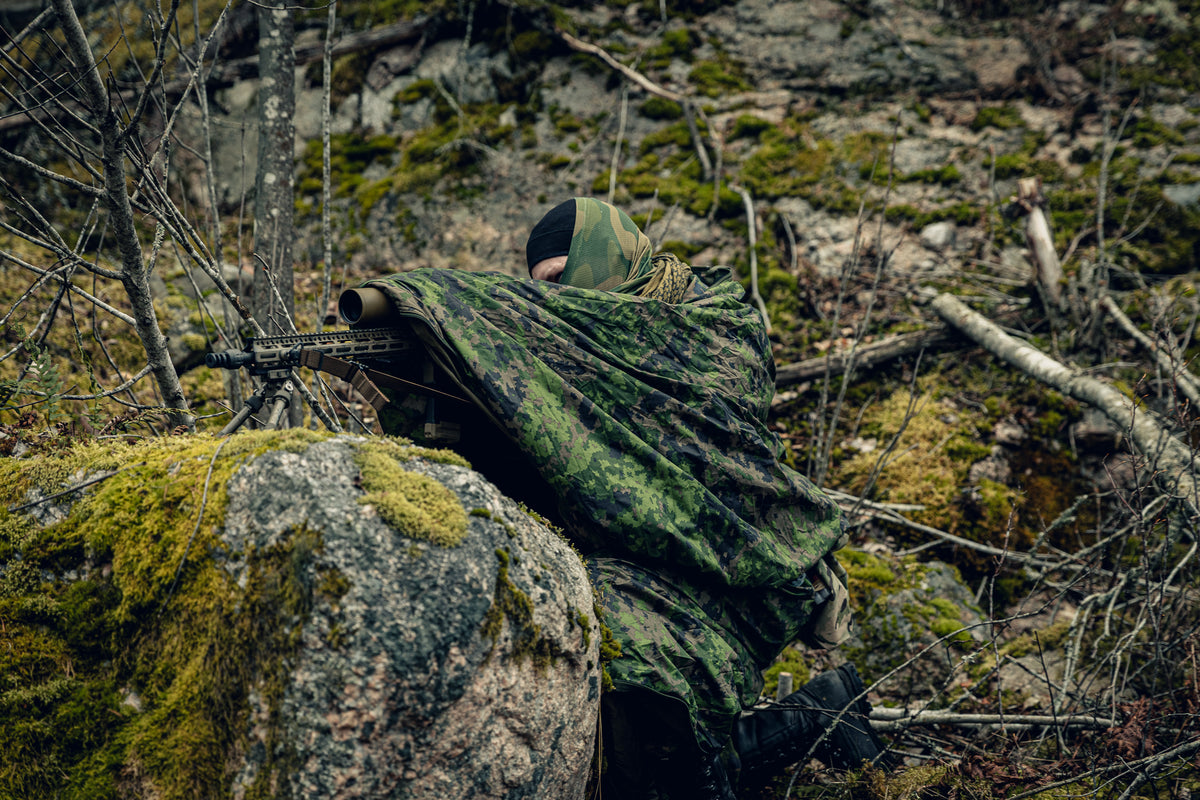 A person kneeling in a forest adjusting a scoped rifle set on a big mossy rock. He is wearing a Finnish M05 woodland cloak, some camouflaged neck tube and a black beanie.