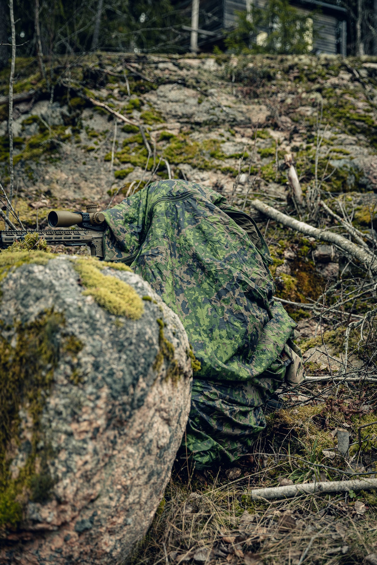 A person in a rocky forest, wearing a cloak in Finnish M05 woodland camo, with a brown scoped rifle coming from underneath the cloak. The rifle is set on a mossy rock, and you can see a small structure in the background.