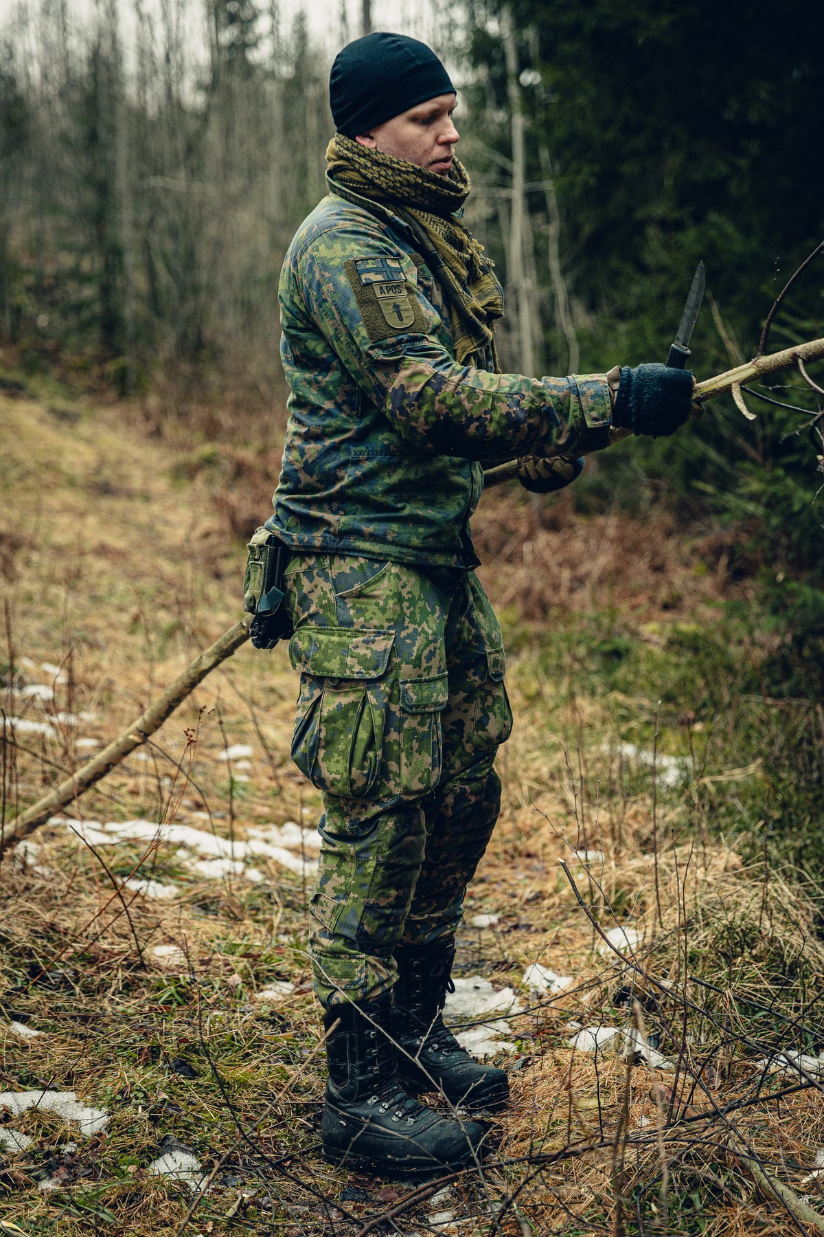 Ein Mann in einem Wald mit einem Ast in der einen und einem Messer in der anderen Hand. Er trägt finnische M05-Tarnbekleidung, eine schwarze Beanie und ein Shemagh-Tuch, und auf seinem Ärmel befinden sich Aufnäher mit der finnischen Flagge, Blutgruppe und Dienstgradabzeichen der Militärpolizei.