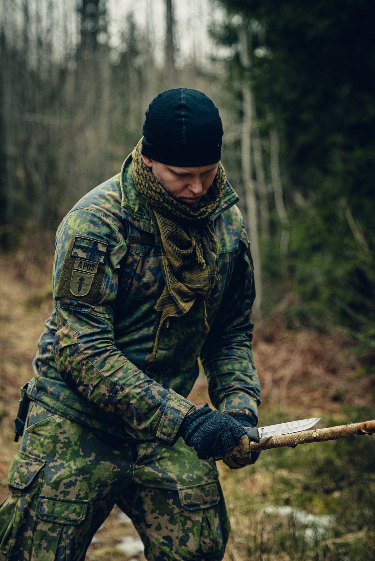 A man in a forest with a tree branch in one hand and a knife in the other one. He is dressed in Finnish M05 camouflage clothing, a black beanie and a shemagh scarf and on his sleeve there are patches with the Finnish flag, blood type and trainint bach insignia for military police.