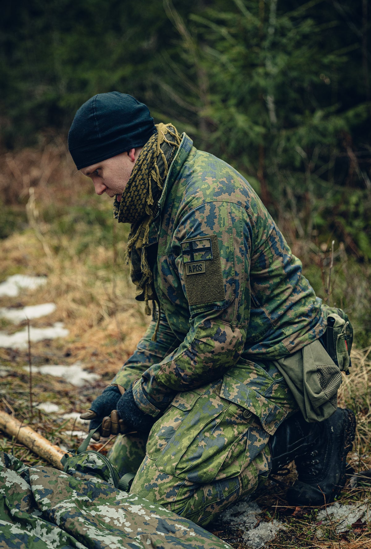 A young man kneeling on the ground in a forest working on attaching a strap to a tree branch, with a M05 winter camo carment lying on the ground. He is dressed in Finnish M05 camouflage clothing, a black beanie and a shemagh scarf and on his sleeve there are patches with the Finnish flag and blood type.
