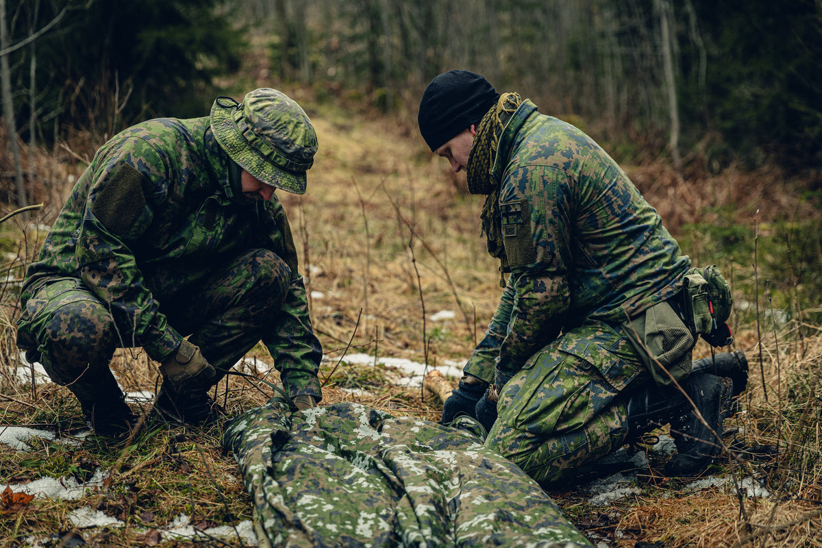 Two men kneeling on the ground fiddling with some sort of garment in Finnish M05 winter camo on the ground. Both are wearing Finnish M05 woodland uniforms, the other one is wearing a M05 boonie hat and the other one a black beanie. 