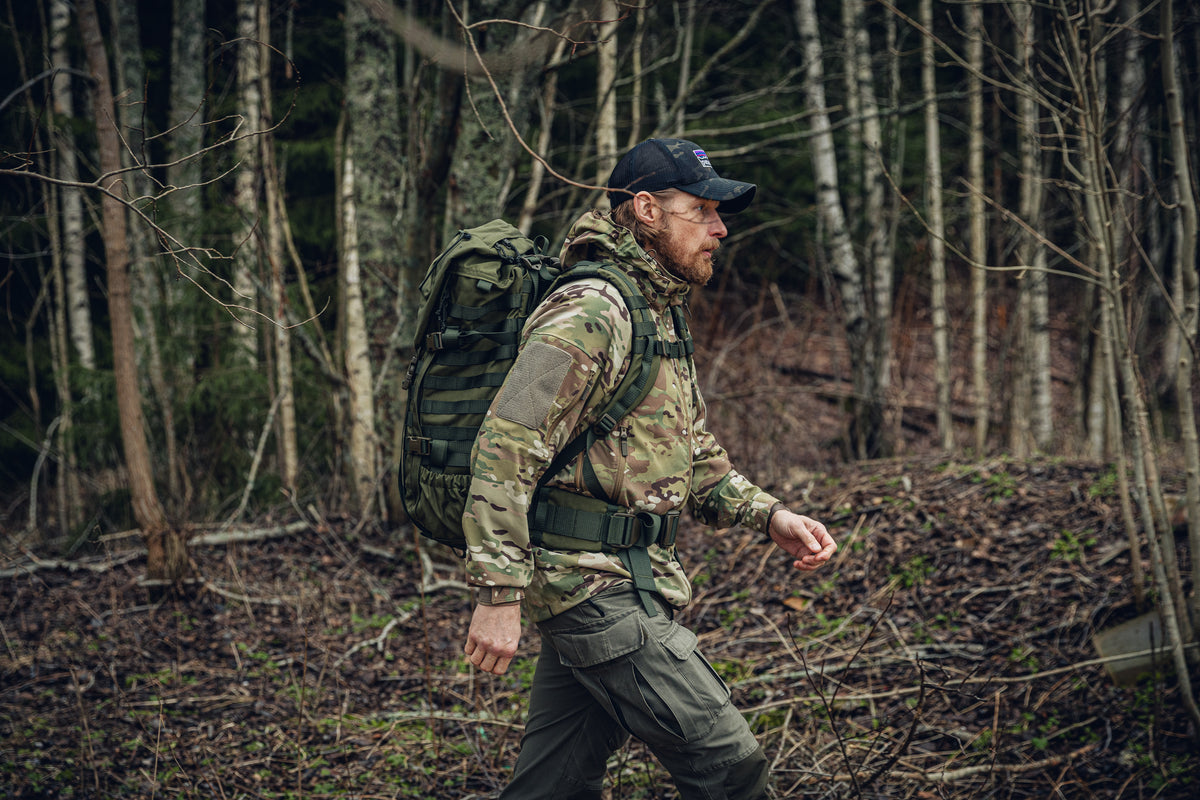A man walking in a forest with a big green backpack. He is wearing green cargo pants, a black camo cap and a multicam jacket.