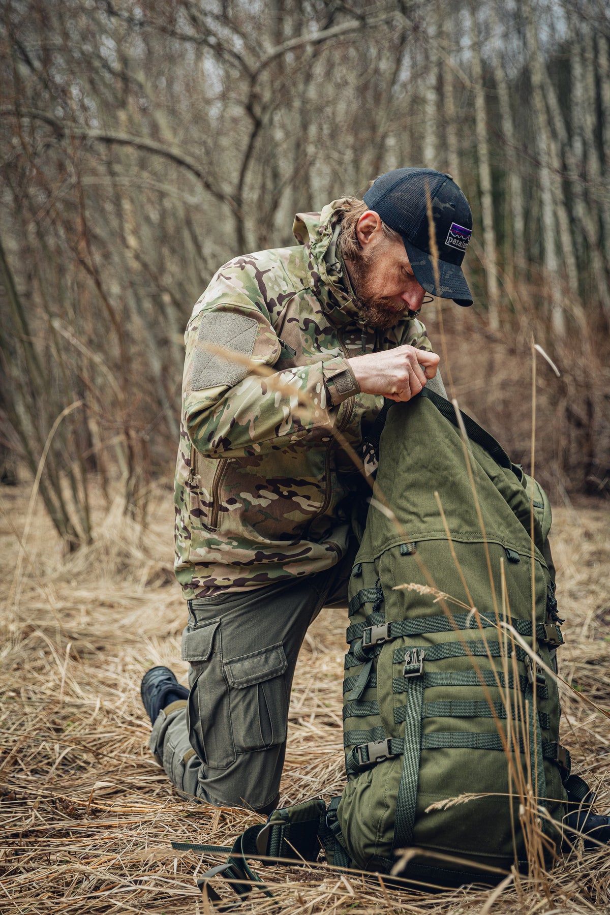 A man kneeling on the ground in a field with some forest in the background, digging through a green backpack. He is wearing green cargo pants and a multicam jacket.