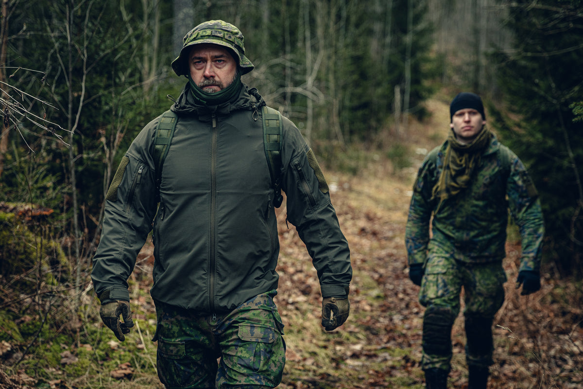 Zwei Männer gehen mit Rucksäcken durch einen Wald. Der eine trägt finnische M05-Waldtarnhosen und einen Boonie Hat, eine grüne Sturmhaube bis zum Kinn heruntergezogen und eine grüne Jacke. Der andere trägt eine komplette finnische M05-Waldtarnuniform und eine schwarze Beanie.
