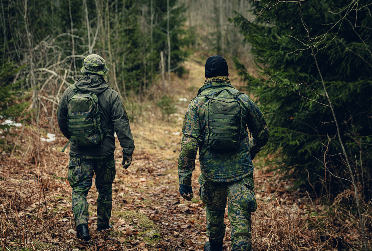 Two men walking away from the camera in a forest. The are wearing Finnish M05 woodland clothing and backpacks.